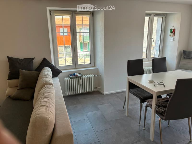 A cozy living and dining area with a beige sofa, a white dining table, and four chairs. The room features large windows with a view of the street and a radiator under the windows.