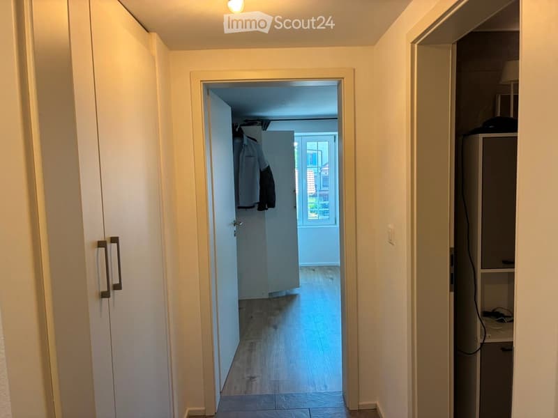 A narrow hallway with light-colored walls and wooden flooring, leading to an open room with a window and some hanging clothes.