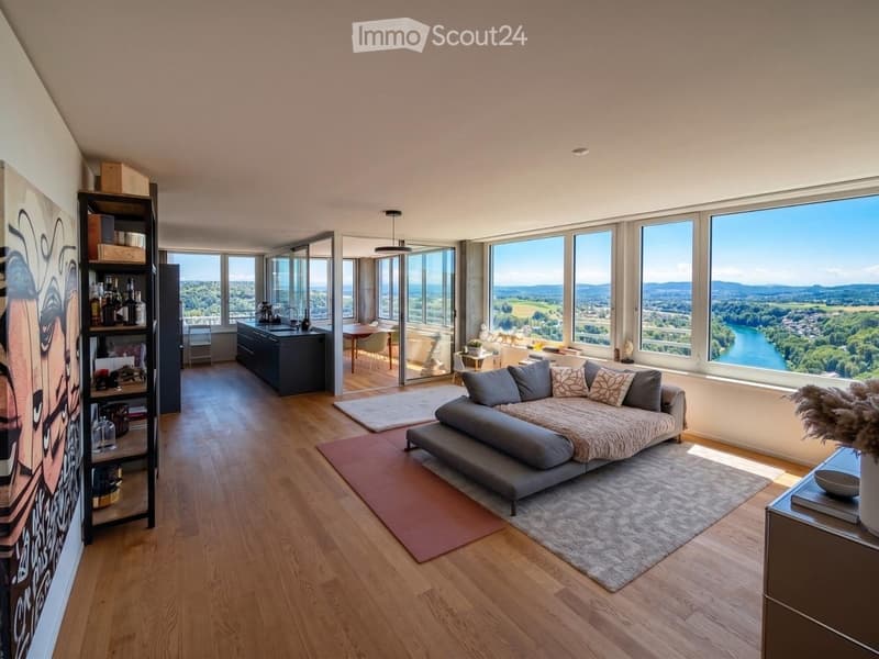 Open-concept living and dining area with large windows offering scenic views, a grey sectional sofa, a coffee table, a bookshelf, a kitchen island, and a dining table with chairs.