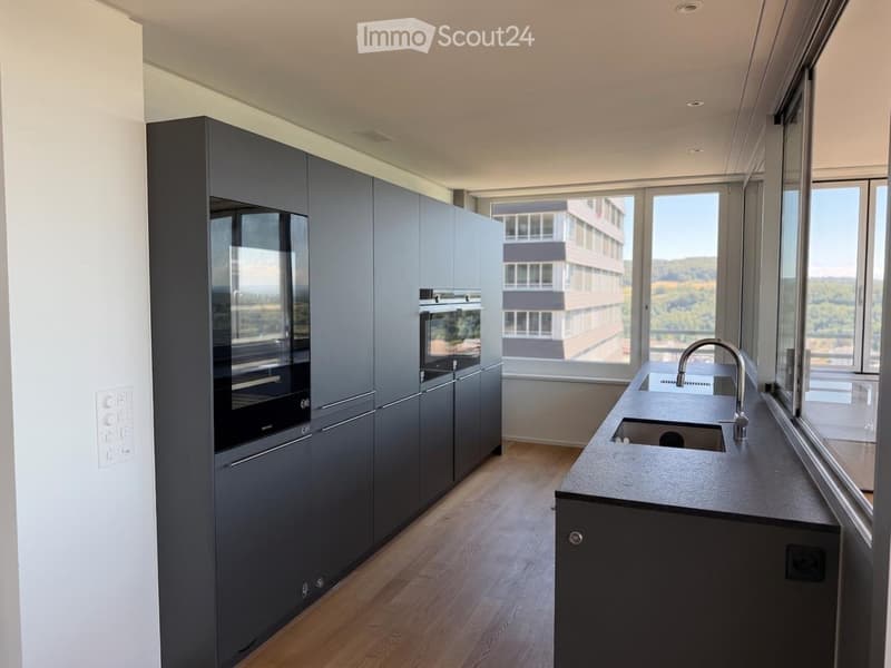 Modern kitchen with dark grey cabinets, a central island with a sink and faucet, and large windows offering a view of the city and landscape.