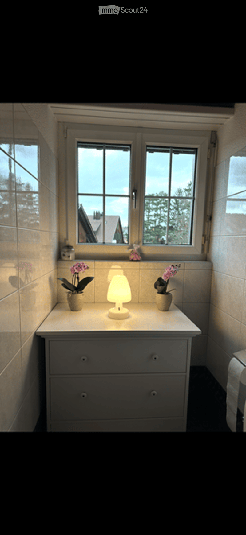 Bathroom corner with a white cabinet, a small table lamp, and two vases with purple flowers on top. The window offers a view of trees and a neighboring house.