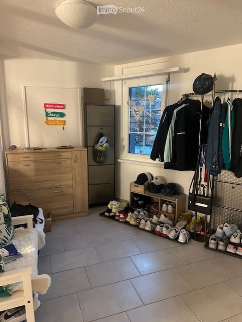 A well-lit hallway or entryway with a sofa, dresser, shoe rack, and a hanger rack. The space features a tiled floor and a window with blinds.