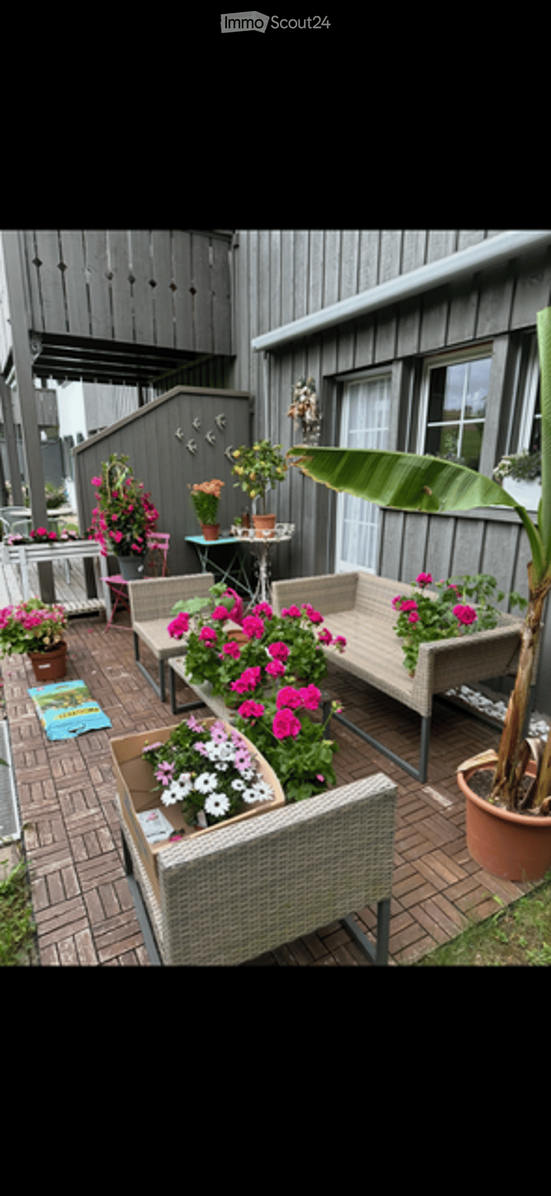 A cozy outdoor patio area with wooden decking, wicker furniture, colorful potted plants, and a small table with chairs.