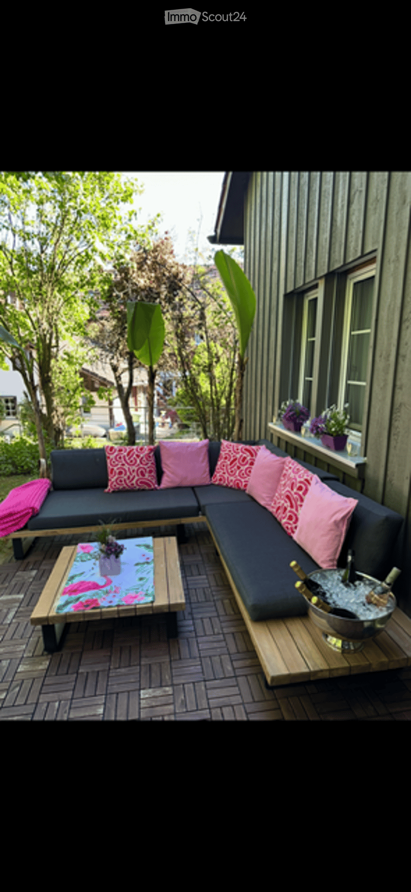 A modern outdoor seating area with a dark grey sectional sofa, pink and red patterned cushions, a wooden coffee table with a floral centerpiece, and a large ice bucket with champagne bottles. The area is surrounded by lush greenery and features a wooden d