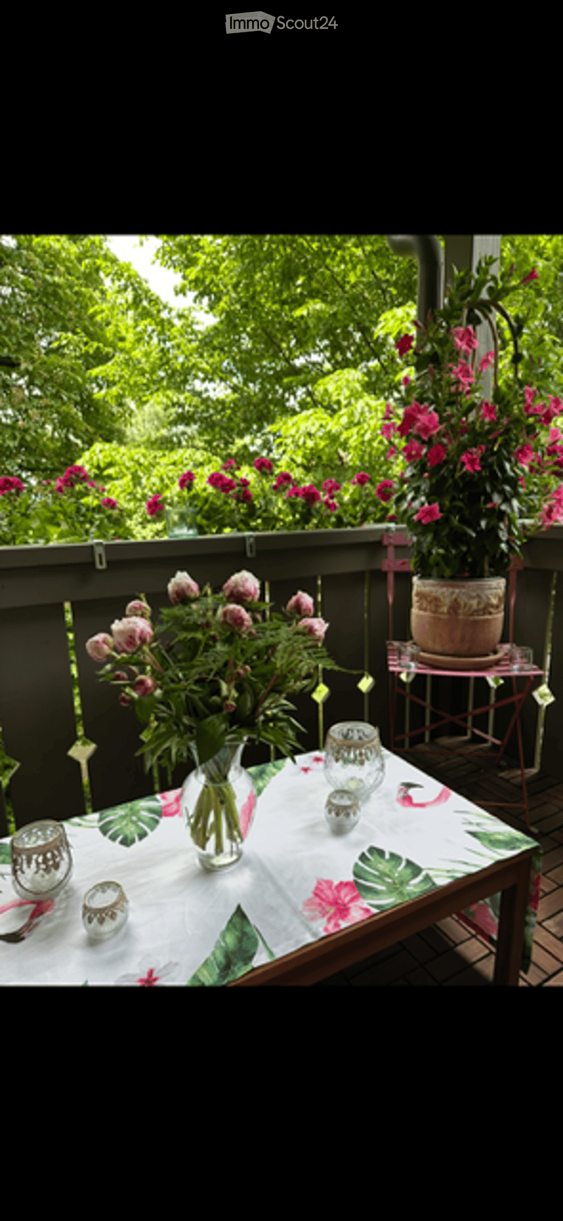 Balcony with a table covered in a floral tablecloth, surrounded by potted plants and flowers, with a railing and greenery in the background.