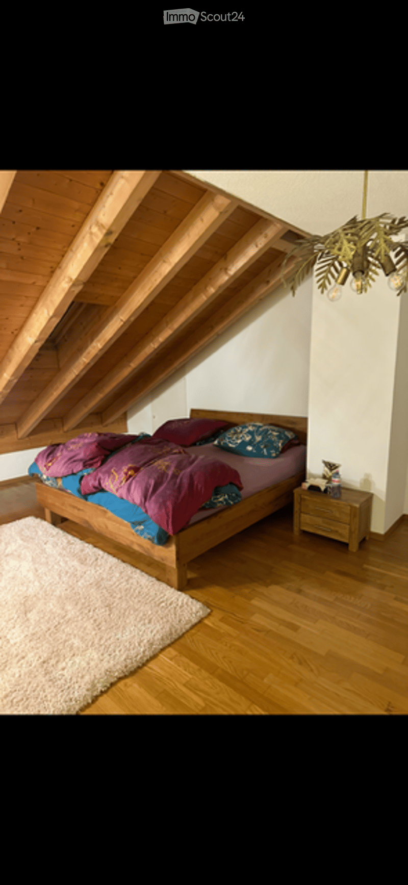 Cozy attic bedroom with wooden slanted ceilings, a large bed with colorful bedding, a nightstand, a fluffy white rug, and a hanging chandelier with a leafy design.