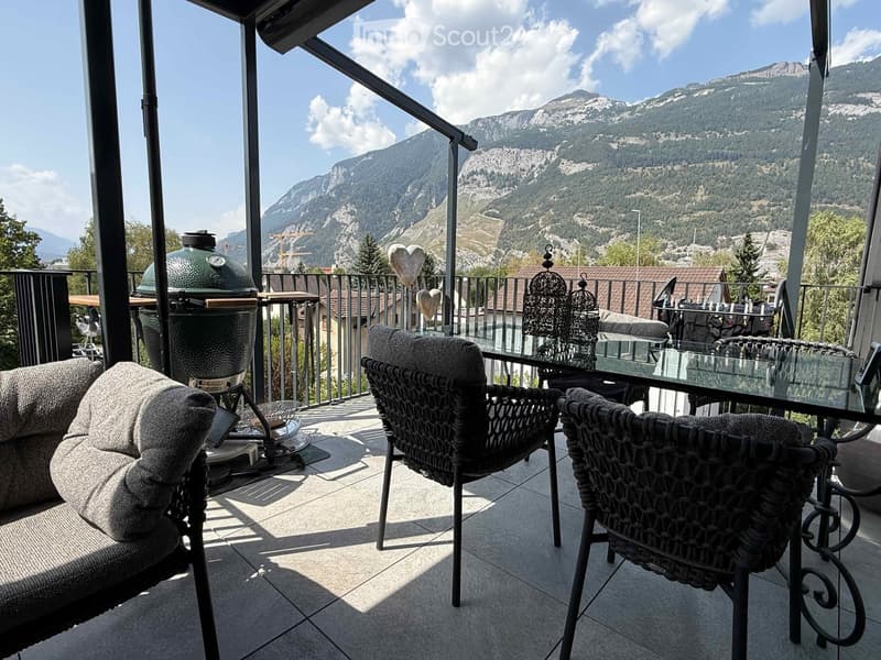 Balcony with a view of mountains, featuring a sofa, chairs, a table, and a grill.