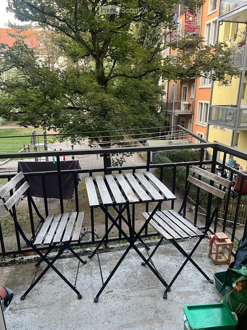 Balcony with wooden folding chairs and a small table, overlooking a residential area with trees and neighboring buildings.