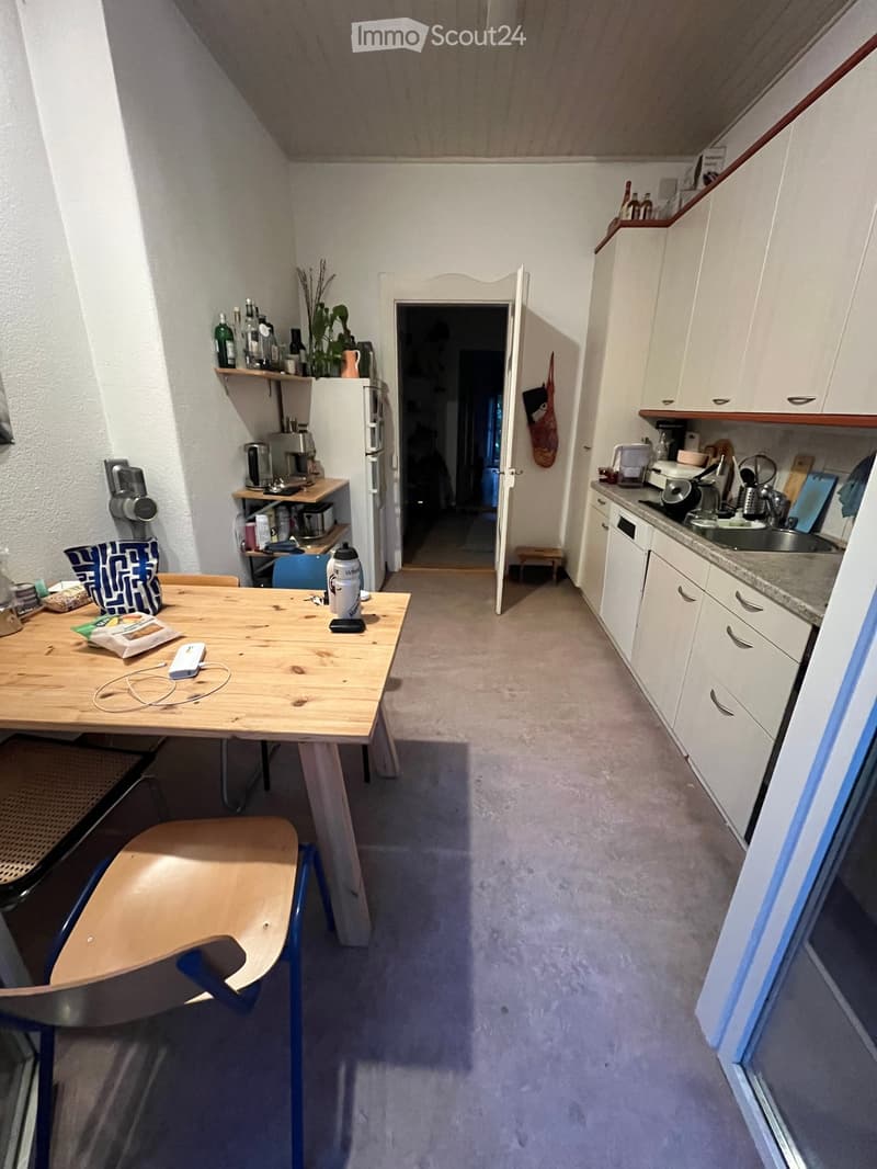 A modern kitchen with white cabinets, a wooden dining table, a chair, a refrigerator, and various appliances including a coffee maker and an oven. The kitchen features a sink and a small stool near the doorway.