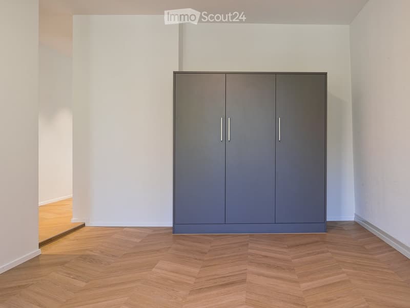 Empty room with light grey walls, wooden herringbone flooring, and a single grey wardrobe with silver handles.