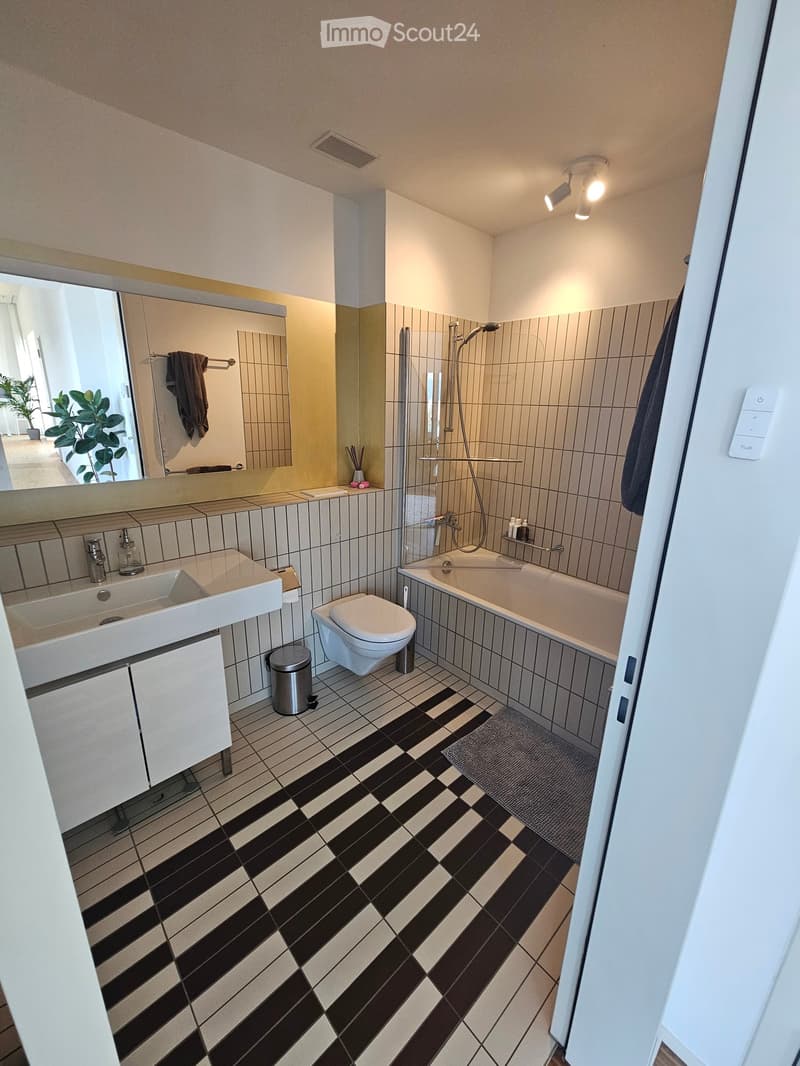 Modern bathroom with black and white tiled floor, white sink, toilet, bathtub, and large mirror. Features a shower area with a glass enclosure and a towel hanging on the wall.