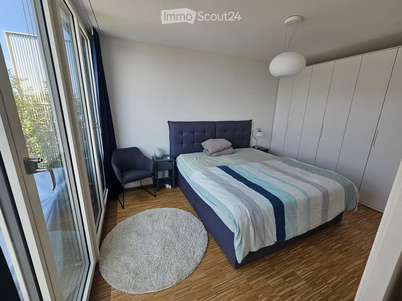 Modern bedroom with a double bed, nightstand, chair, round rug, and built-in wardrobe. Large window with blinds offers natural light.