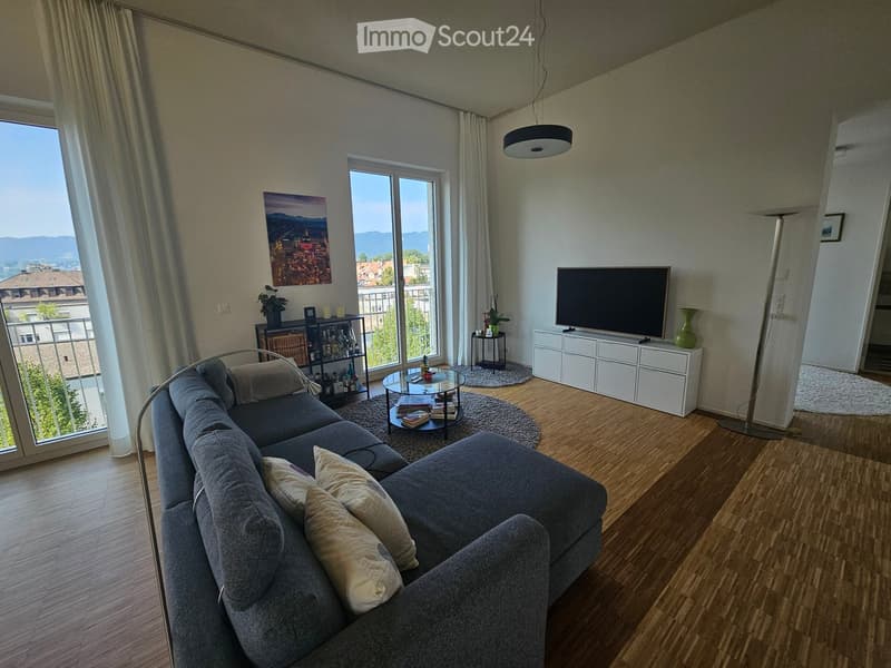 Modern living room with a grey sectional sofa, coffee table, TV stand, floor lamp, side table, shelf, vase, and picture frame. Large windows offer a scenic view.