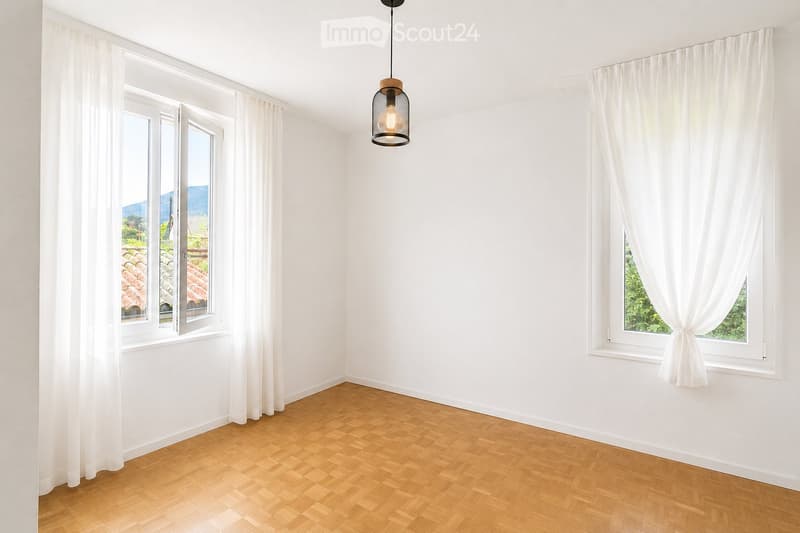 Empty room with wooden flooring, white walls, large windows with white curtains, and a hanging light fixture.