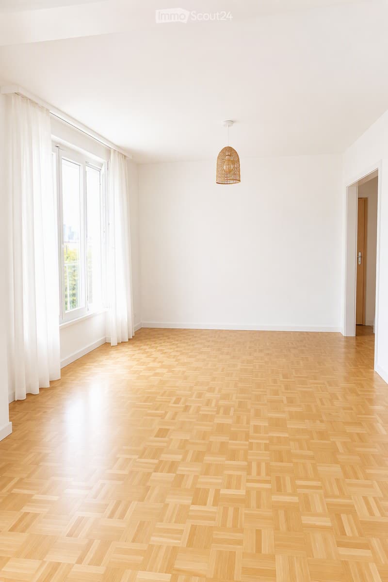 Empty room with light wooden herringbone flooring, white walls, large windows with white curtains, and a single hanging light fixture.