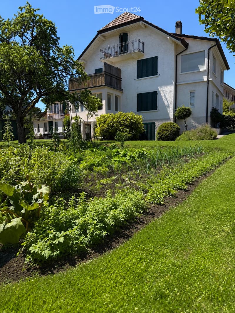 A well-maintained garden with a variety of plants and vegetables, leading up to a two-story white house with green shutters and balconies.