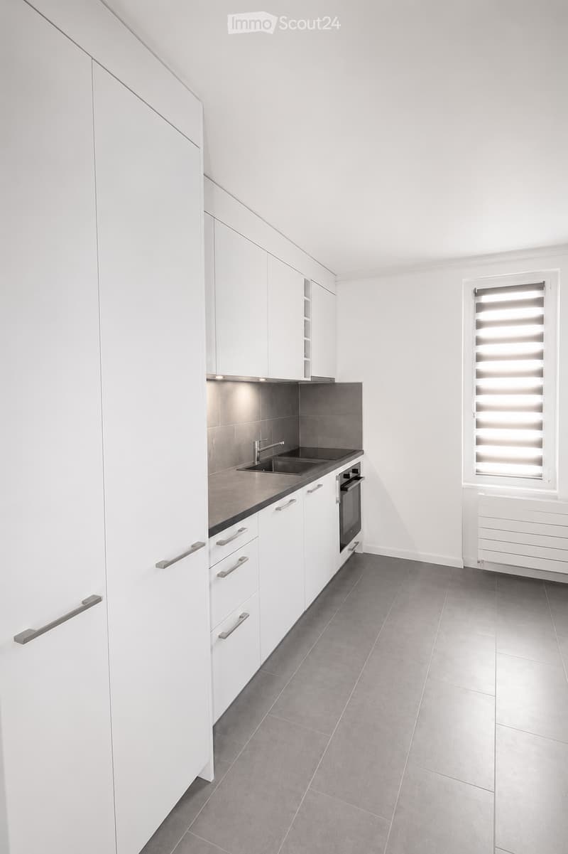 Modern kitchen with white cabinets, stainless steel sink, and built-in oven. Grey tiled floor and walls.