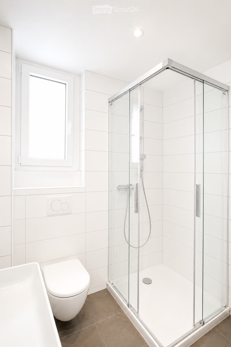 Modern bathroom with white tiles, a bathtub, toilet, and a glass-enclosed shower with a handheld showerhead.
