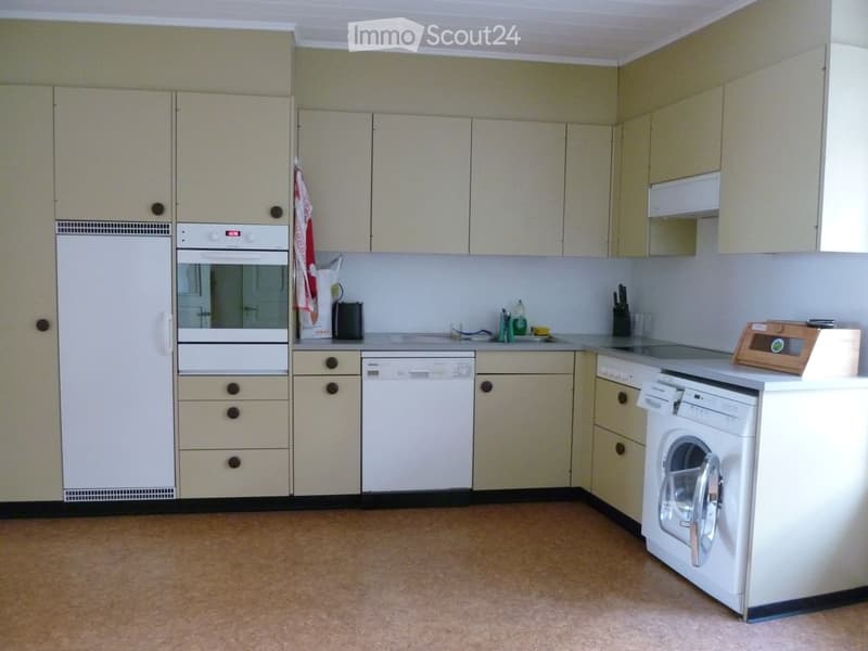 A well-lit kitchen with light-colored cabinets, a refrigerator, oven, dishwasher, and washing machine. The countertop has various small items including a cup, bottle, and knife block.