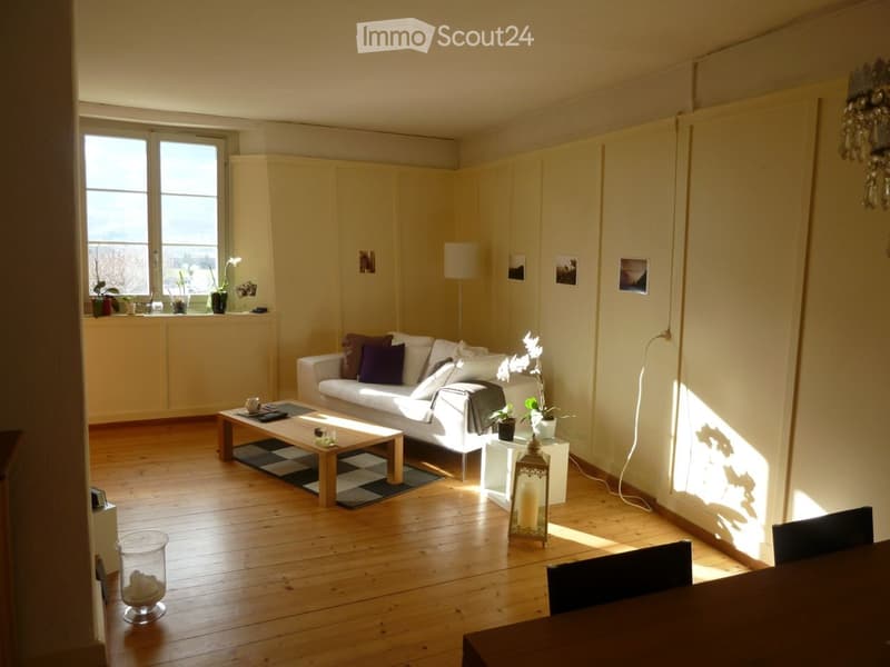Bright living room with wooden flooring, white walls, large windows, a white sofa with colorful cushions, a coffee table, a dining table with chairs, and decorative items including candles and vases.