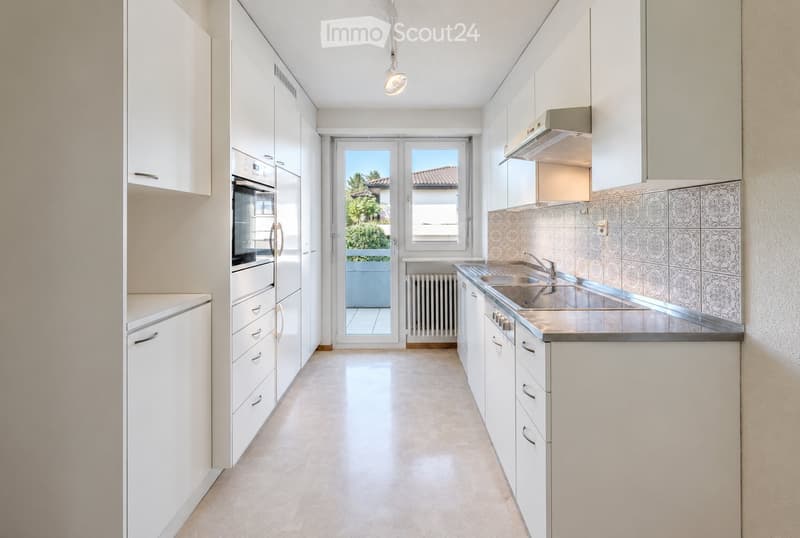 Modern kitchen with white cabinets, stainless steel sink, built-in oven, and a view of a balcony through a glass door.