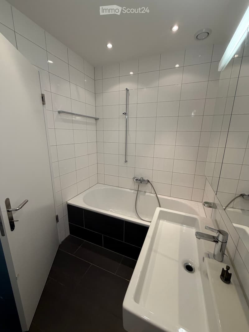 Modern bathroom with white tiled walls, a bathtub, a double sink, and a large mirror. Features include a handheld showerhead, a door with a silver handle, and recessed ceiling lights.