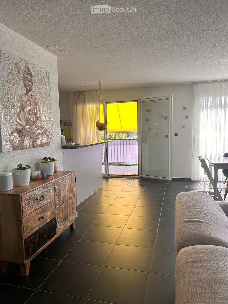 Modern living room with a sofa, coffee table, dining table, chairs, a wooden cabinet, plants, and a large Buddha wall art. The room features a sliding glass door leading to a balcony.