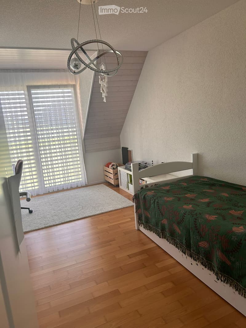 Cozy bedroom with wooden flooring, slanted ceiling, large window with blinds, bed with green patterned blanket, small table, chair, and decorative lamp.