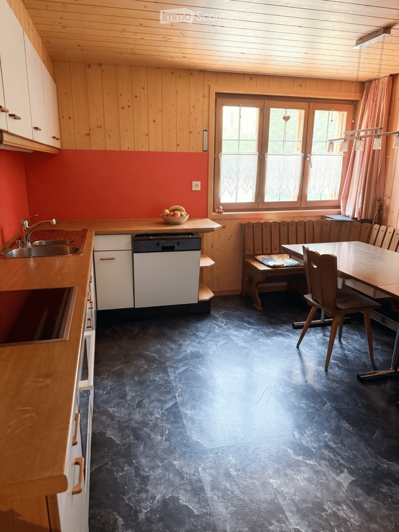 Kitchen with wooden cabinets, a sink, dishwasher, and a dining area with a table and chairs. The floor is dark blue, and there is a bowl of fruit on the counter.