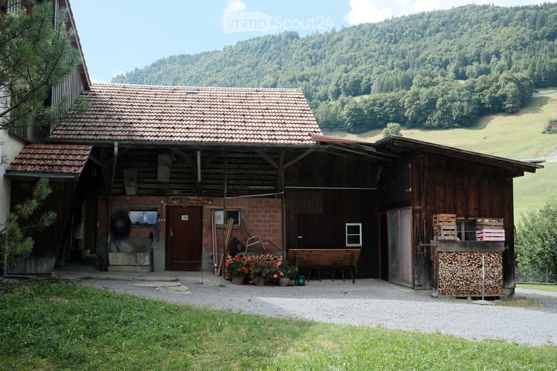 A rustic wooden barn with a tiled roof, brick walls, and a large open garage area. The barn features a wooden workbench, potted flowers, a stack of firewood, and a tire on the left side.