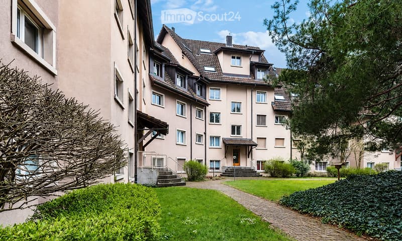 A multi-story residential building with a beige facade, multiple windows, and a well-maintained garden area with green lawns and trimmed bushes.