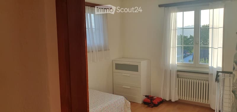 Bedroom with a bed, chest of drawers, radiator, and a drill on the floor. The room has wooden flooring, white walls, and two windows with white curtains.