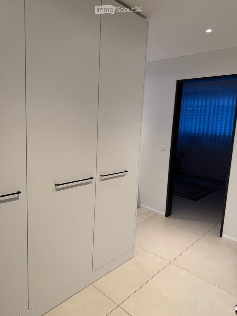 A modern hallway with light-colored tiled flooring, white walls, and a large mirrored closet with silver handles. A doorway leads to another room with a glimpse of a dark-colored rug and part of a piece of furniture.