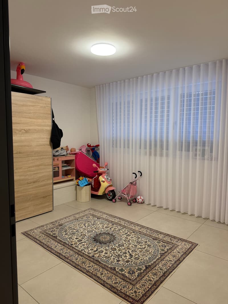 A well-lit room with a large window covered by white curtains, a patterned rug on the floor, a wooden wardrobe, a pink scooter, a baby walker, and various toys.