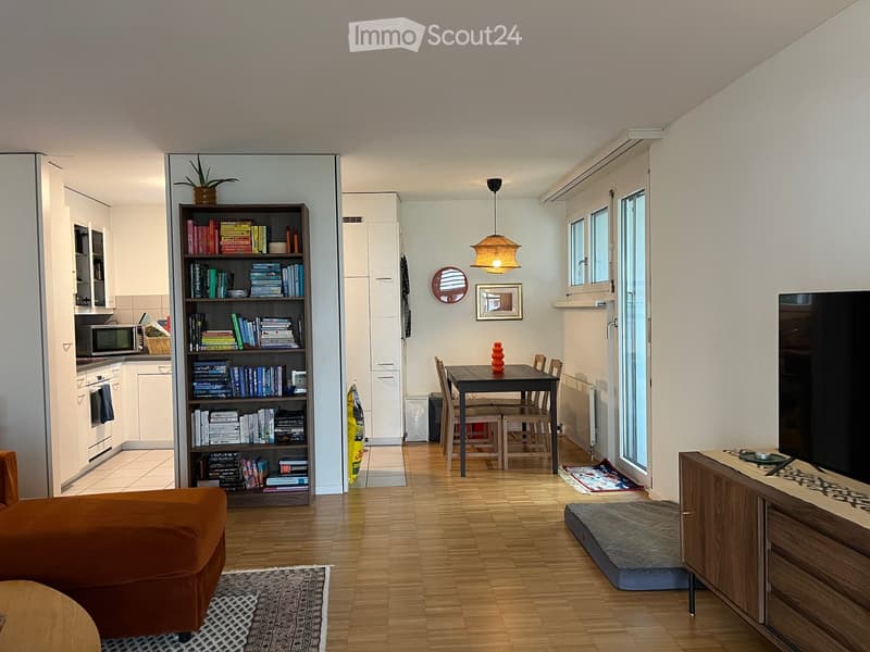 A modern, open-concept living and dining area with wooden flooring, a bookshelf filled with books, a sofa, a dining table with chairs, a kitchen area with appliances, and a TV stand with a screen.