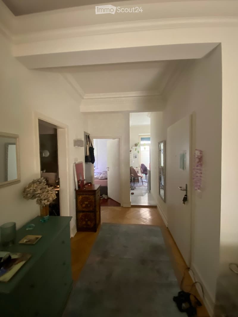 A well-lit hallway with wooden flooring, white walls, and several doors leading to different rooms. There is a small rug in the center, a green side table with a vase of flowers, and a decorative cabinet with ornate carvings.