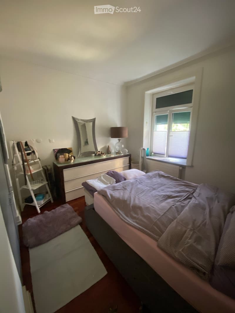 Cozy bedroom with a double bed, nightstand, mirror, lamp, and a small shelf. The room features wooden flooring and a window with blinds.