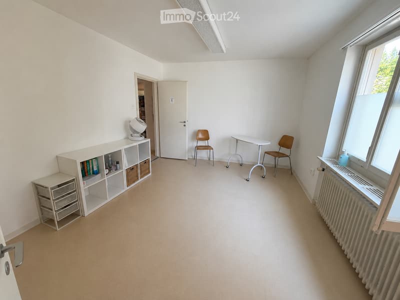 Bright, unfurnished room with large windows, a small table, two chairs, a shelf with books and storage boxes, and a fan on top of the shelf.