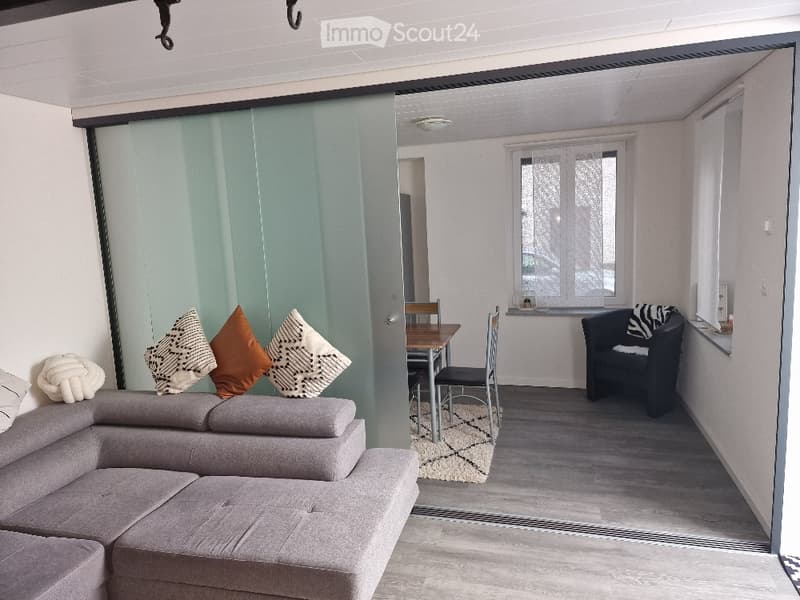 Modern living room with a grey sectional sofa, colorful throw pillows, a small dining area with a table and chairs, and a black armchair near a window.