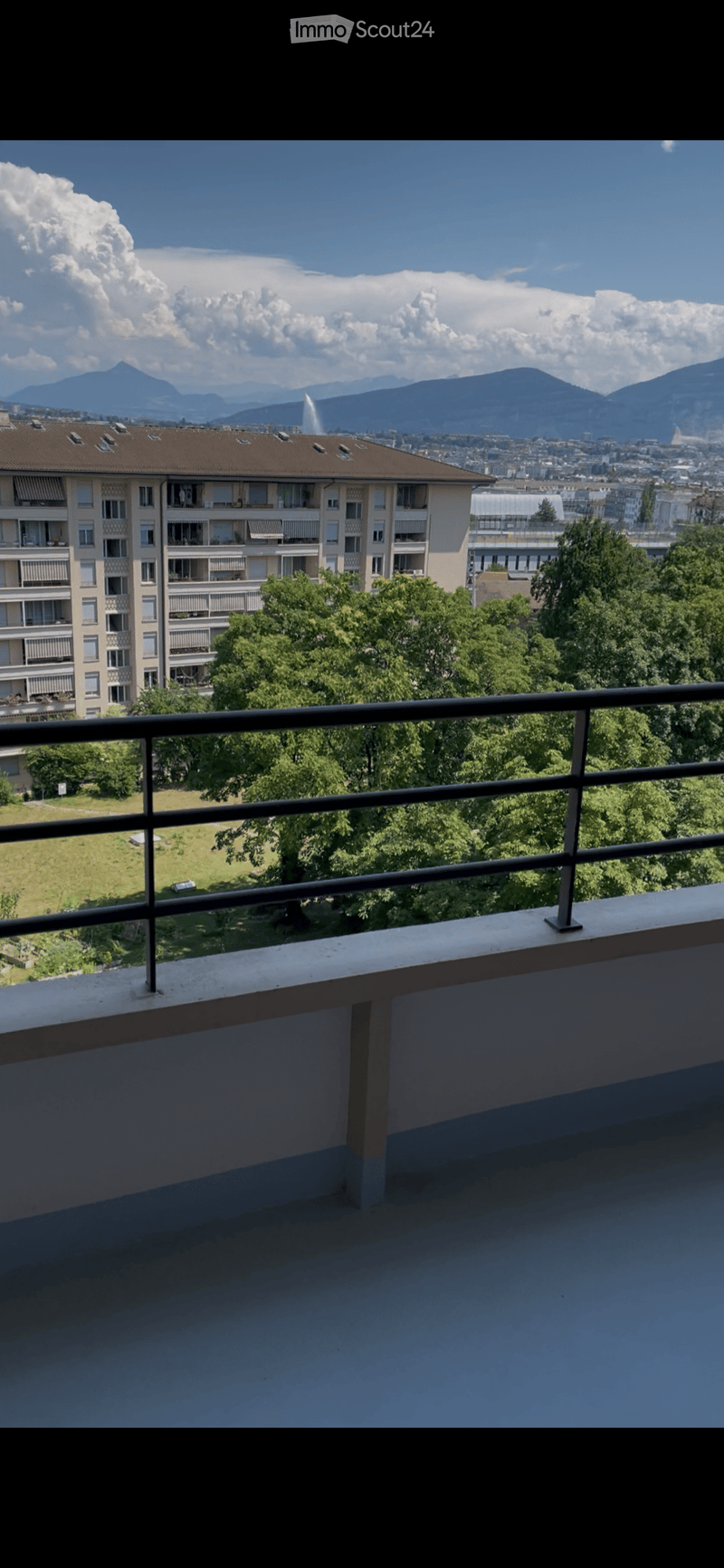 Balcony with metal railing, overlooking a park and cityscape with mountains in the background.