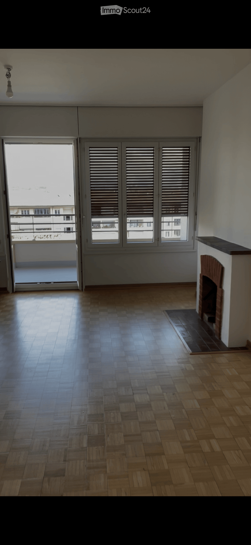 Empty room with wooden flooring, fireplace, large windows with shutters, and a balcony view.