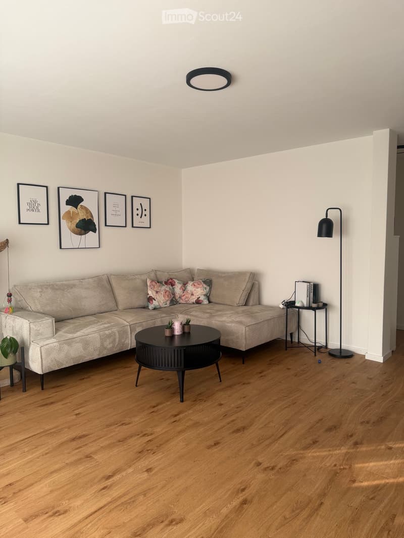 Modern living room with light-colored sectional sofa, coffee table, floor lamp, side table, potted plants, and picture frames on the wall.