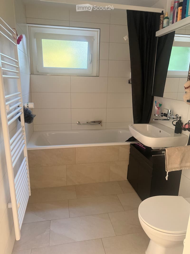 Modern bathroom with a bathtub, sink, toilet, and a radiator. Features tiled walls and floor, a window with frosted glass, and a black shower curtain.