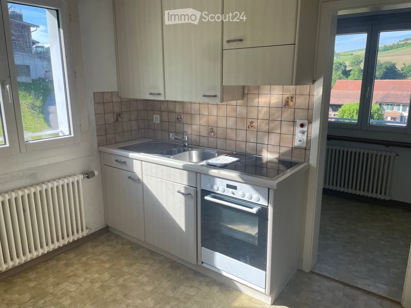 Modern kitchen with light wood cabinets, stainless steel sink and oven, tiled backsplash, and large windows offering a view of the countryside.