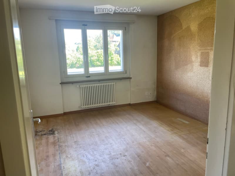 Empty room with wooden flooring, white walls, a window with white frames, and a radiator below the window.