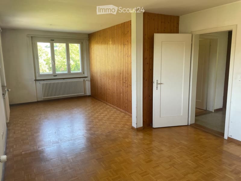 Empty room with wooden flooring, white walls, and a large window with a radiator below it. The room has wooden paneling on one wall and a white door leading to another space.