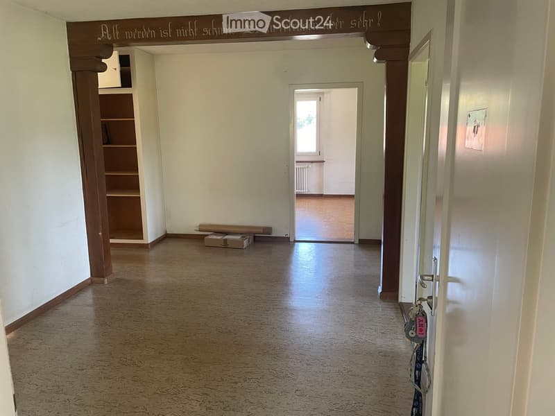 Empty hallway with light-colored walls, wooden floor, and a small shelf on the left side. A doorway leads to another room with a window and radiator visible.