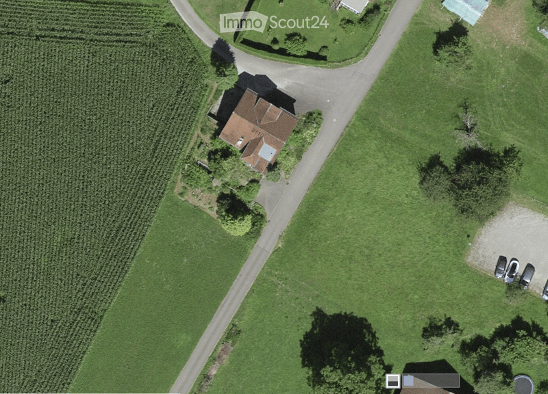 Aerial view of a single-story house with a brown roof, surrounded by green fields and a driveway leading to a parking area with several cars.