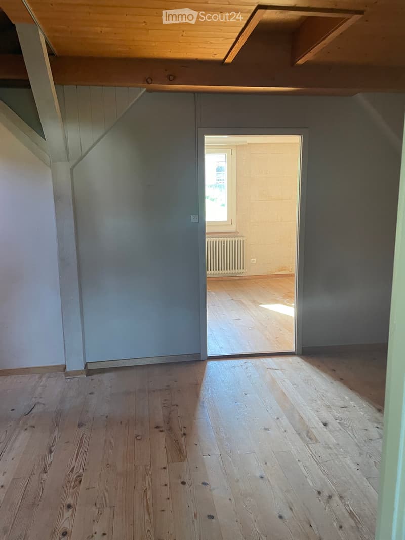 Empty room with wooden flooring and exposed wooden beams on the ceiling. The room has grey walls and a doorway leading to another room with a radiator and a window.
