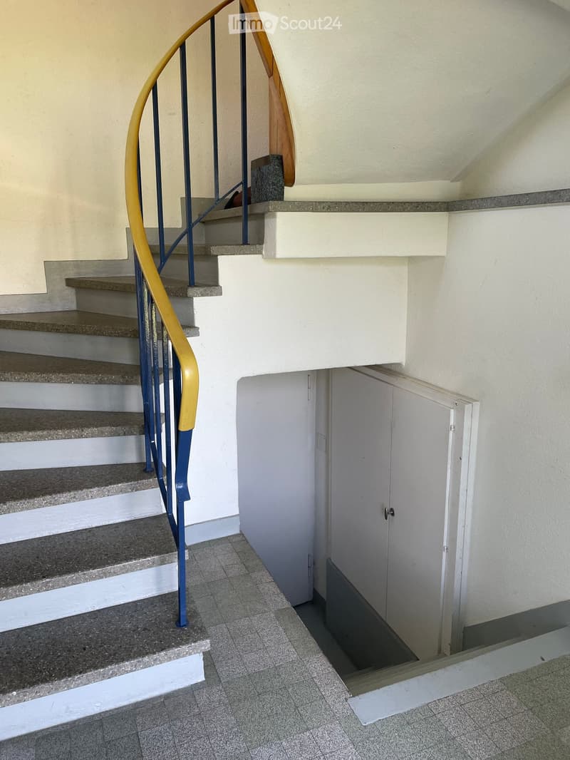 Staircase with blue and yellow handrail, gray tiled floor, white walls, and a closed white door leading to another area.